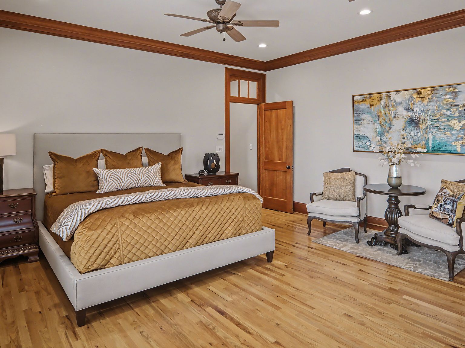 A primary bedroom with quilted bed, sitting area, and large gold-framed artwork above warm oak floors